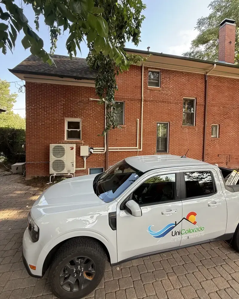 A UniColorado pickup parked beside a newly installed heat pump on the side of a red-brick house.